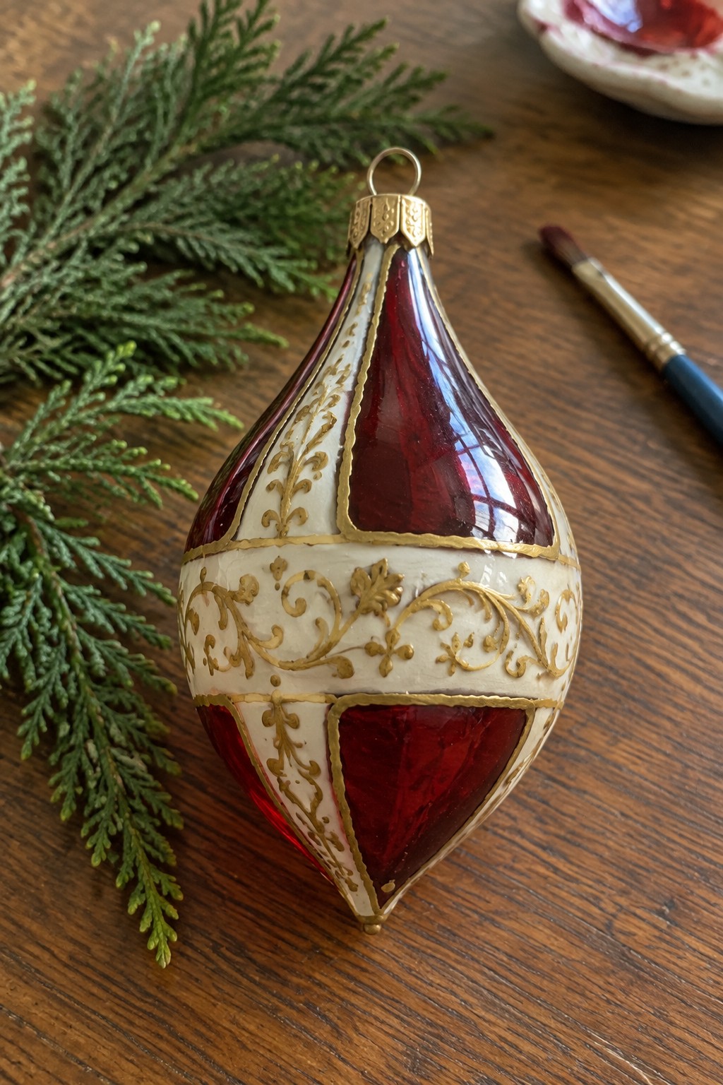 Ornate red glass teardrop ornament with gold filigree beside evergreen branches and paintbrush on wood table
