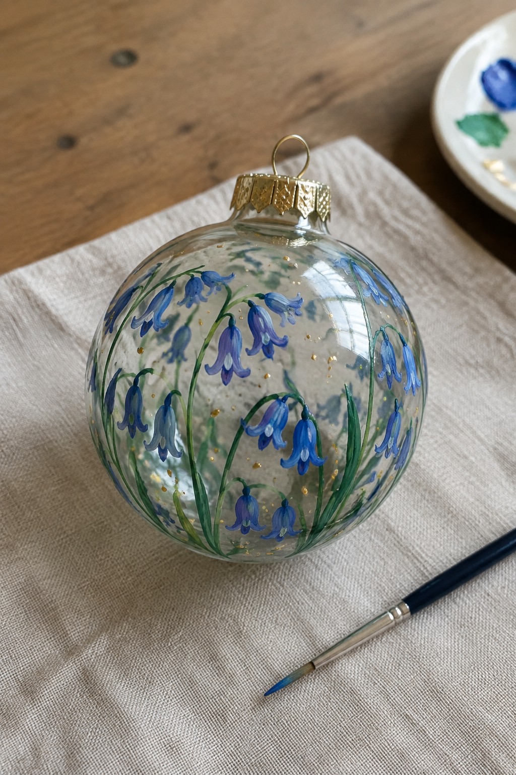A clear glass spherical ornament painted with bluebell flowers and gold dots rests on a linen cloth beside a paintbrush.