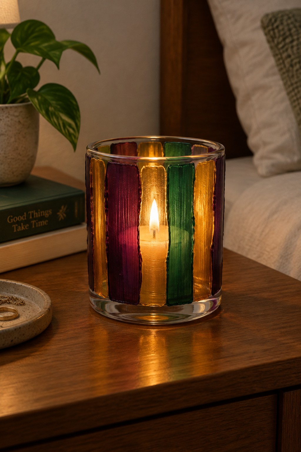 Lit candle in vibrant striped glass holder on wooden nightstand with plant and book.