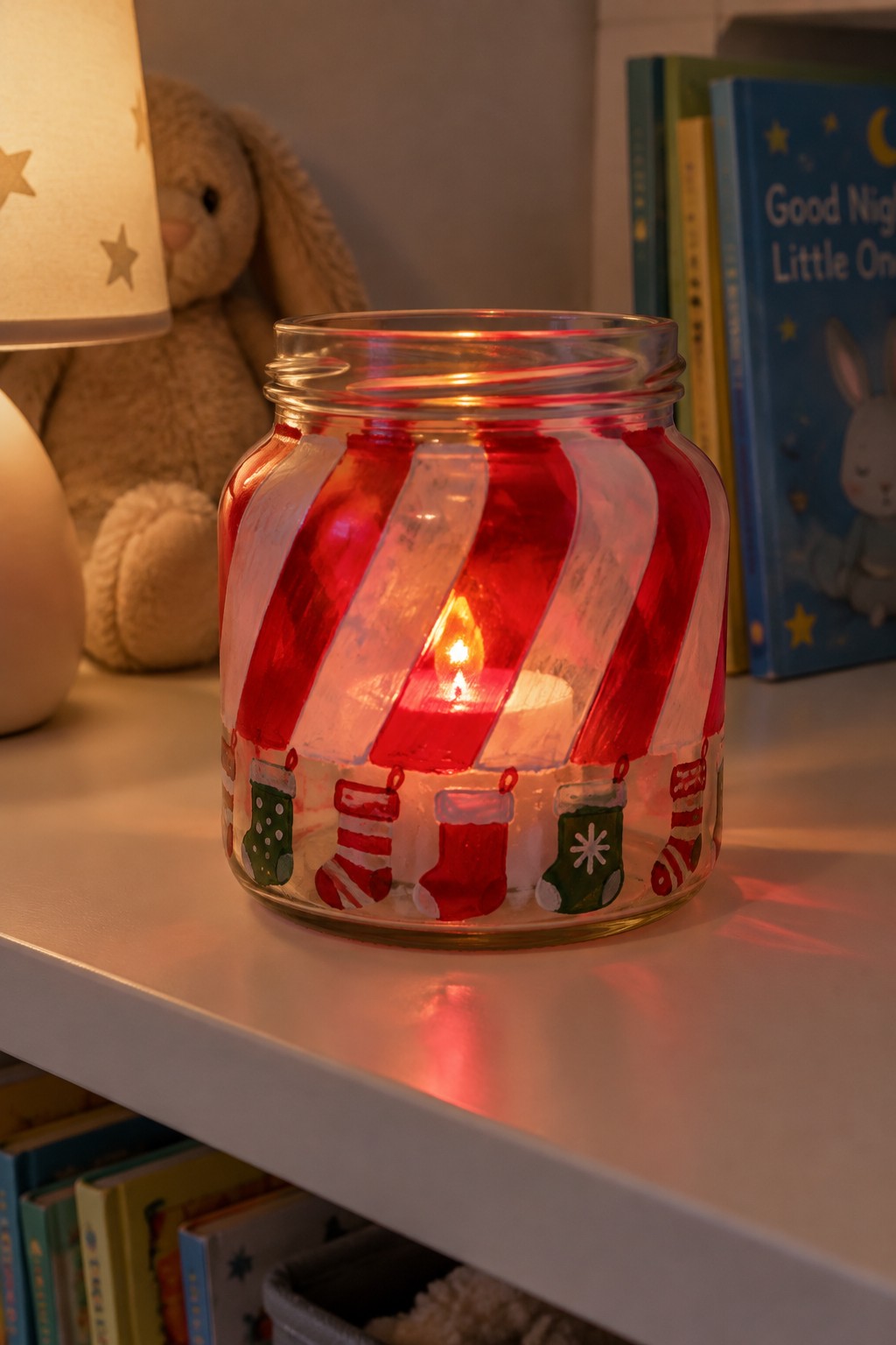 Lit candle in red-striped glass jar with painted Christmas stockings on shelf